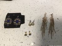 Four pairs of earrings displayed on a white surface, including one pair of large purple stone chandeliers, long chain tassel earrings, and two pairs of small stud earrings.