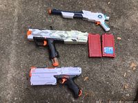 Photo showing three Nerf guns arranged on the ground: one white and gray camouflaged rifle with black handle, one white rifle with teal and orange accents, and one black and white pistol along with two red Nerf dart magazines.
