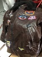Front view of brown leather jacket showing multiple colorful military patches and zippered front closure.