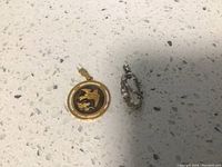 Two charms on flat surface, one gold plated with black and bird design, other silver-tone with clear stones.