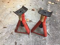 Two red metal jack stands placed side by side on a concrete floor. Both show visible surface rust and worn paper labels on the vertical support arm. Each has a black metal saddle top and a triangular base for stability.