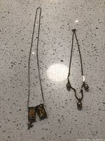 Two vintage necklaces displayed on a white stone surface, one long with religious rectangular pendants and cross, the other short with small pearl-like beads and religious charms.
