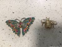 Two vintage pins placed on a speckled countertop: one butterfly-shaped pin with multicolored stones and one bee-shaped pin with clear stones.
