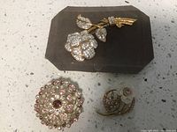 Three vintage pins with stone embellishments displayed on a black velvet cushion; one large flower, one circular, and one crescent floral pin.