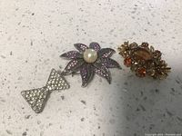 Three vintage pins on white speckled surface: silver-tone bow pin, flower pin with purple stones and pearl, gold-tone floral pin with brown and orange stones.