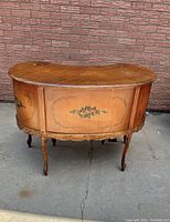 Front view showing kidney-shaped form and hand-painted green floral accents on wood panels with cabriole legs and carved apron.