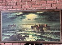 Full view of the framed vintage textured lithograph showing a moonlit ocean with boats and rocky foreground, set against a brick wall.