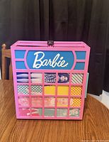 Front view of pink Barbie carry case with white Barbie logo and grid compartments showing dolls inside.