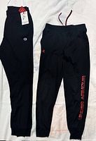 Two pairs of black boys pants side by side on white background. Left: Champion sweatpants with tags. Right: Under Armour leggings with red logo and lettering.
