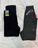 Black Tek Gear jogging pants and gray Champion Granite Heat jogging pants laid flat on surface showing full length and detail tags