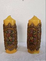 Front view of both yellow vintage pillar candles with decorative floral relief designs in multicolor and ornate frame patterns.