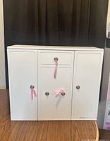 Front view of white doll closet with closed doors, metal knobs, and pink ribbons.
