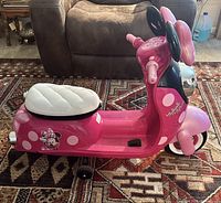 Side profile of pink Minnie Mouse themed ride-on scooter showing white seat, black training wheels, and side Minnie Mouse decal.