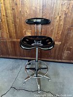 Front view of black draft swivel chair with adjustable height and chrome metal base with footrest ring, placed on concrete floor in front of wood paneled wall.
