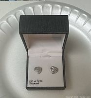 Heart-shaped diamond stud earrings displayed in a black jewelry box on a white surface.