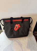Front view of black Rolling Stone tote bag showing the classic red tongue logo and red zipper detail