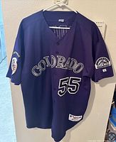 Front view of navy blue Colorado Rockies baseball jersey with number 55 and 'COLORADO' text. Hanging on a hanger against wall.