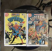 Two vintage Superboy comic books in protective sleeves displayed on a stool; one issue is #1 from April 1979 with yellow cover and Superboy fighting thugs, the other is issue #2 from May 1979 with an action scene on cover.