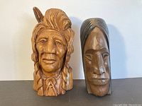 Photo of two wooden First Nations bust carvings side by side on gray surface and white background. The busts show detailed faces and hair styling.