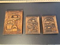 Three carved cedar plaques with West Coast designs on a black surface showing overall lot