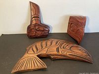 Overview of three carved cedar pieces on black surface: salmon, whale and red-stained panel.