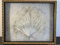 Full view of antique feather fan mounted in a glass-covered shadow box with ornate gold frame and textured background.