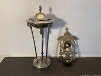 Wide view of the silver ciborium with lid and porcelain miniature of Christ with metal frame side by side on a dark surface.