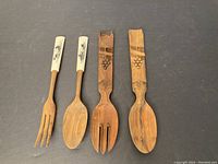 Photo of two pairs of vintage serving utensils on black background: one pair wooden with carved grape cluster handle motif, other pair wood with ceramic tops decorated with black botanical designs.