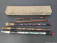 Full view of five vintage calligraphy brushes alongside a vintage patterned storage box, showing variety in bristle type and handle decoration.