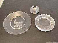 Three glass items on a black surface: Lalique dish with frosted fern pattern, Val Saint Lambert plate with etched rabbit, antique glass candle holder with silver insert