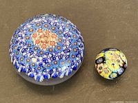 Photo of two mille fleur paperweights side-by-side on a flat surface. Larger one predominantly blue flowers, smaller with yellow base.