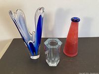 Three art glass vases displayed on table with blue and clear flowing vase, clear geometric vase, and red frosted Moretti vase.