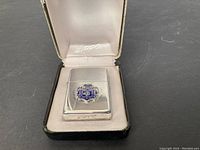 Front view of the sterling silver Zippo lighter with blue and silver crest emblem inside white satin-lined black presentation box stamped with Zippo.