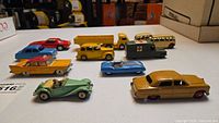 Overview of ten vintage diecast toy cars including sedans, buses, trucks, ambulance, and sports cars in various colors placed on a white surface.