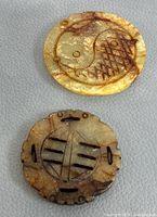 Close-up photo of two round hand carved pendants on a light grey surface, showing the yin-yang design and geometric carving with brown and yellow tones.