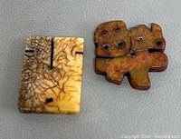 Photo of two hand carved Chinese stone/jade pendants on a gray surface. One pendant is a stylized Chinese character in mottled brown and yellow stone with drilled holes. The other is a rectangular pale brown pendant with a crackled surface and incised lines.