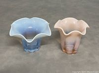 Two vintage Duncan and Miller opalescent glass vases, blue and pink, with ruffled edges, placed side by side on neutral surface.