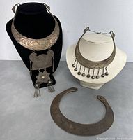 Three handmade old tribe ceremonial metal collar necklaces displayed on stands, showing the varying sizes, detailed etching, pendants and dangling elements.