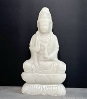 Front view of white stone Buddha seated on lotus base against black background.