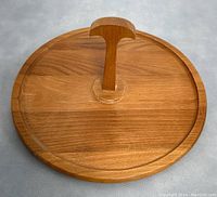 Front view of teak wood round cheese tray with central handle, showing the wood grain, curved handle, and a dividing groove on the surface.