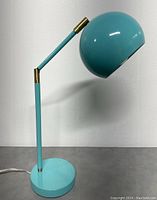 Turquoise desk lamp with adjustable arm and dome-shaped shade. Side profile showing the lamp in an upright position with brass accent at the joint.
