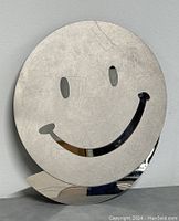 Front angled view of the heavy metal smiley face wall decor showing its reflective surface and some wear marks.