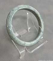 Natural green jade bangle displayed on clear stand, showing the round shape and color.