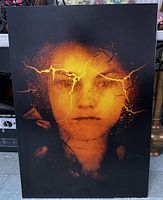 Front view of the metal print showing the surreal cracked face portrait in orange and black tones