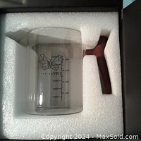 Top-down view of the clear glass pouring pot with a dark wooden handle, resting in white foam inside a black box.