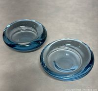 Pair of blue and clear glass bowls with thick rounded rims placed on flat surface. Bowls are hollow and signed by designer.