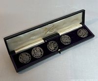 Five hallmarked Art Nouveau style silver buttons arranged in original black box with purple velvet lining and cream satin lid interior, showing good condition and detailed design.