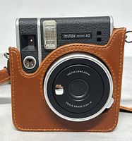 Front of Fujifilm Instax Mini 40 instant camera in brown leather protective case, showing lens, flash, and body details.