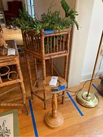 Bamboo hexagon plant stand with plants inside, standing next to a smaller turned wood pedestal stand and a brass floor lamp on hardwood floor.