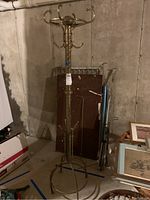 Full view of one brass coat and hat rack standing in basement against concrete wall. Surrounded by miscellaneous items such as framed pictures and boxes.